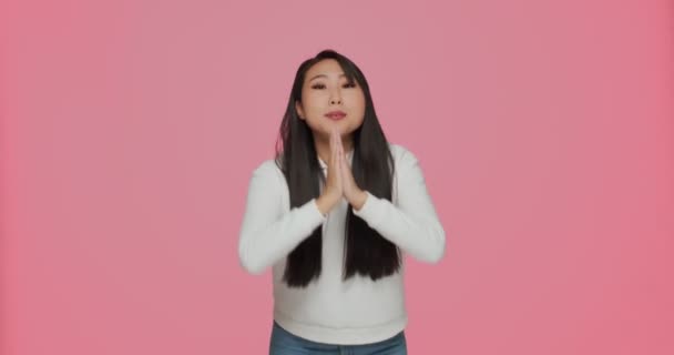 Азійська молода жінка, бажаючи чогось складеними руками, просить допомоги або прощення, просячи на рожевому фоні студії — стокове відео