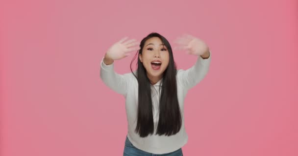 Happy friendly asian young girl waving hands in greeting gesture looking at camera, welcoming on pink studio background — Vídeo de Stock