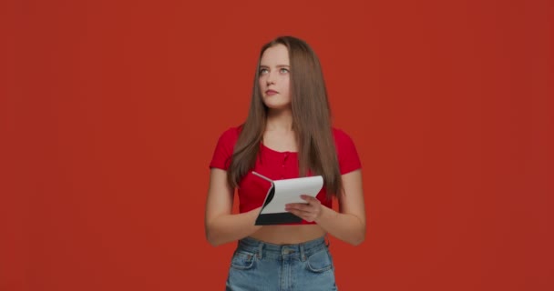 Menina moderna Pensive escrever notas no bloco de notas, inventar novas ideias, planejamento, fazer a lista em fundo laranja — Vídeo de Stock