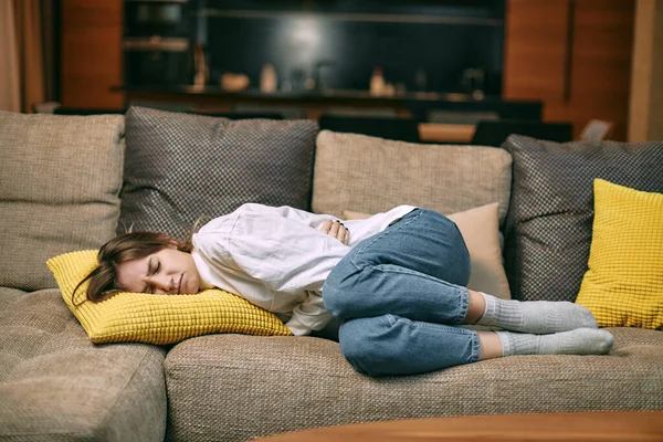 Unhealthy young woman suffering menstrual pain, holding belly, having abdominal cramps during period lying on couch — Zdjęcie stockowe