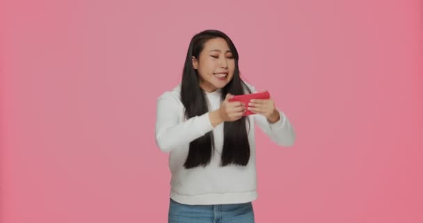 Funny asian girl using mobile phone with attentive focused expression playing video game on cellphone on pink background — Stockvideo