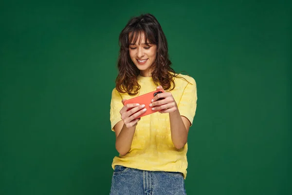 Happy modern young girl using mobile phone application playing video game on cellphone on green studio background — Stock fotografie