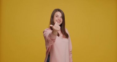 Happy young girl make beckoning hands gesture, asking come here, inviting, welcoming, smiling on yellow background