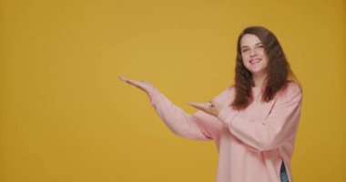 Cheerful young girl showing empty copy space by palms, advertise product presenting service on yellow studio background