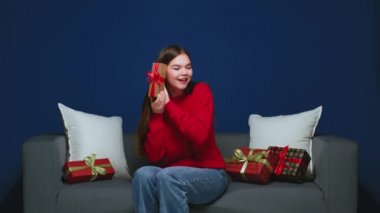 Happy young girl shaking wrapped gift box, guessing what is inside, sitting on couch. Festive surprise concept
