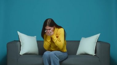 Ill allergic young girl sneezing in tissue, blowing running nose, suffering flu cold or allergy symptom sitting on couch