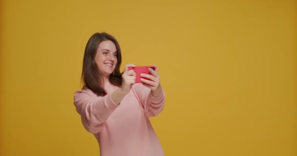 जुआ युवा लड़की पीले स्टूडियो पृष्ठभूमि पर आधुनिक मोबाइल अनुप्रयोग का उपयोग करके फोन पर वीडियो गेम खेल रही है — स्टॉक वीडियो