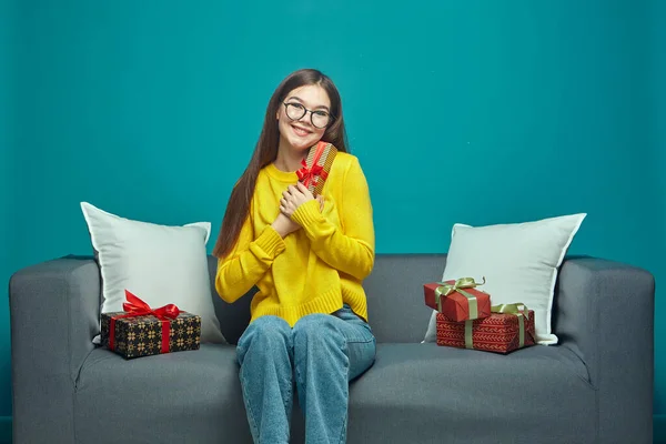 Joyful young girl in glasses holding festive gift sit on couch with wrapped holiday gifts boxes, celebratory presents — Stockfoto