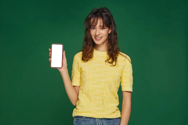 Smiling beautiful young girl show phone with blank mock-up screen, advertising new apps on green studio background — Stock fotografie