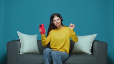 Dancing young modern girl in headphones cool moving seated on sofa, enjoying favorite music using phone application