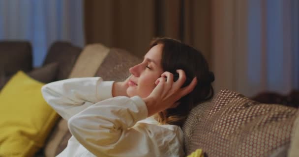 Calm young woman put on modern wireless headphones to relax on comfortable sofa, listening to music, stress-free concept — Stock video