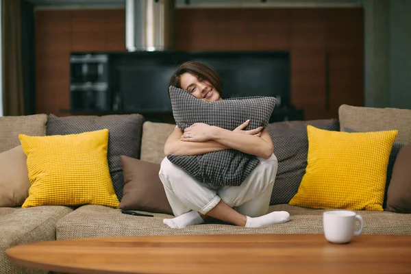 Happy young woman smiling hugging pillow sitting on sofa in living room at cozy home, resting, relaxing on weekend — Stok fotoğraf