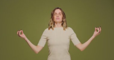 Young woman calming, breathing deep, meditating with mudra om gesture by hands, doing yogs exercise on green background