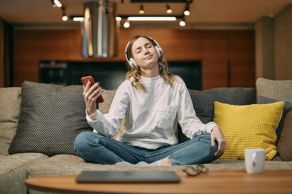 Young woman in headphones enjoying relaxing music, using phone with closed eyes resting on comfortable couch at home