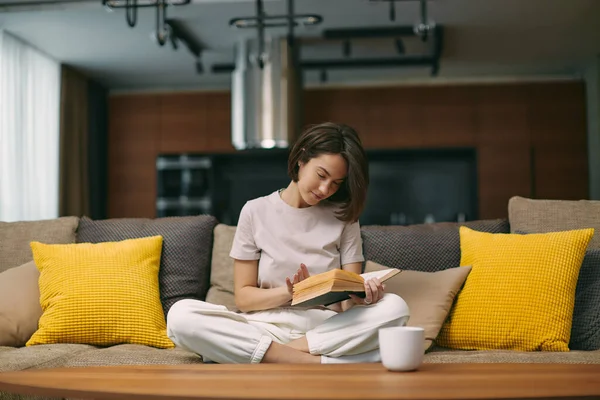 Modern young girl reading bestseller novel in paper book, enjoying weekend, relaxing on sofa at home. Self-education — Stok fotoğraf