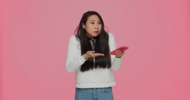 Puzzled confused young asian girl reading spam message holding phone, shrug shoulders on pink studio background