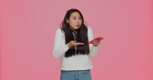 Puzzled confused young asian girl reading spam message holding phone, shrug shoulders on pink studio background — Stockvideo