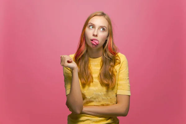 Funny young girl showing tongue with foolish face, fooling, making faces on pink background. Playful childish mood — Stockfoto