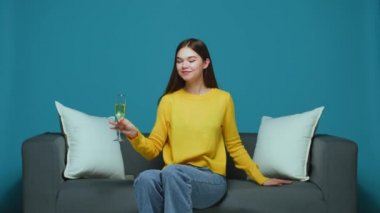Smiling young girl clink by glass of sparkling wine, drink fizz champagne sitting on sofa, celebrating holiday party