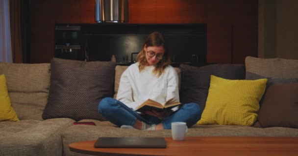 Молода жінка читає цікаву книгу, сидячи на дивані, насолоджується вільним часом вдома. Самоосвіта, бестселерівська література — стокове відео