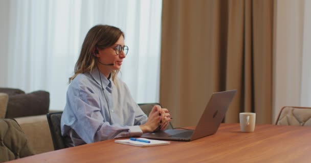 Vriendelijke jonge vrouwelijke manager in headset, werken aan laptop in gesprek met partners of klanten online. Afstandsbediening — Stockvideo