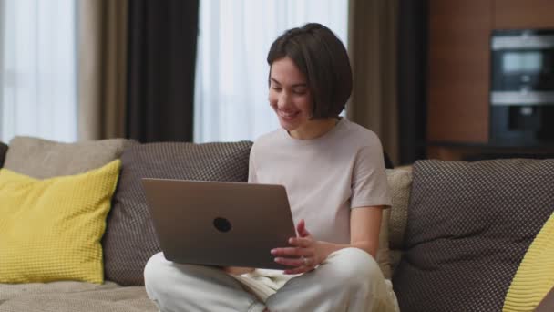 Позитивна молода дівчина сидить за допомогою ноутбука махаючи рукою до камери привітання, прощання на віддаленому відео уроці, сидячи на дивані вдома — стокове відео