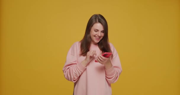 SNSで携帯電話を持っている幸せな若い女の子が黄色い背景で楽しそうに笑っています。出会い系アプリ広告 — ストック動画
