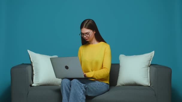 Poważna nowoczesna dziewczyna w okularach siedząca na kanapie z laptopem, pracująca lub ucząca się online, pisząca na komputerze — Wideo stockowe