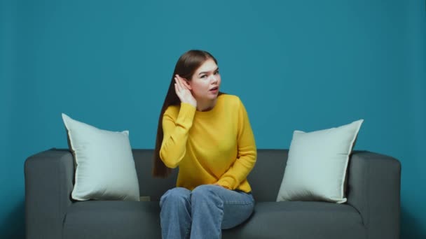 Nowoczesna młoda dziewczyna trzymająca się za rękę blisko ucha słuchająca uważnie, mająca problemy ze słuchem, pytająca co, siedząca na kanapie — Wideo stockowe
