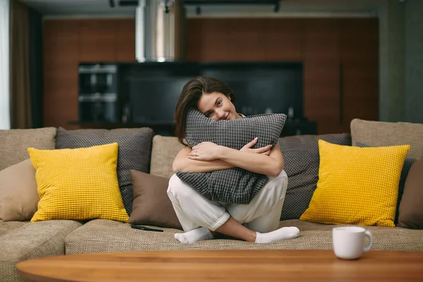 Mutlu genç kız rahat koltukta oturan yumuşak yastığa sarılıyor. Rahatlamış kadın evdeki modern oturma odasında dinleniyor. — Stok fotoğraf