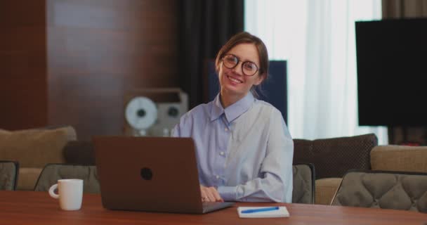 Glimlachende jonge studente in bril zit achter een bureau met laptop, kijkend naar de camera thuis. Online onderwijs, e-learning — Stockvideo