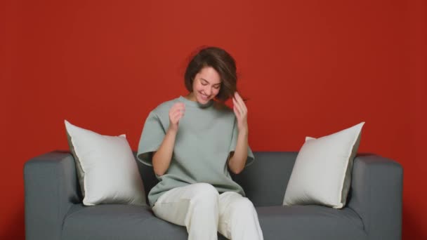 Una joven con un hermoso peinado está tonteando, jugando con su pelo, agitando la cabeza mientras está sentada en el sofá en casa. — Vídeos de Stock