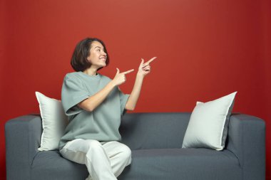 Uzayı kopyala Gülümseyen modern genç kadın boş bir yeri işaret ediyor, kırmızı stüdyo arka planında koltukta oturuyor. İyi bir teklif.