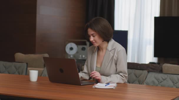 Професійна усміхнена молода жінка-бізнесмен, що сидить за столом, працює на ноутбуці з даними, друкує електронну пошту, п'є чай — стокове відео