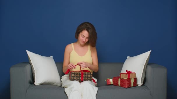 Egy fiatal anya karácsonyi ajándékokat pakol a családjának. Egy masnit köt egy ünnepi szalagra. Ajándékokat pakol a kanapéra.. — Stock videók