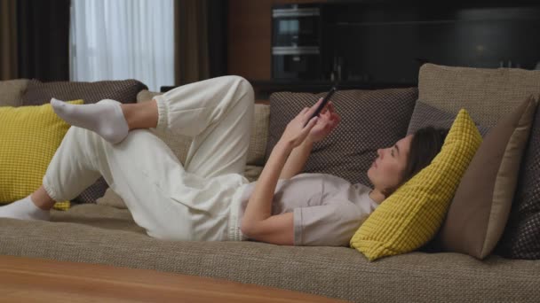 Young girl using modern phone applications lying on comfortable couch at home shopping, looking social networks — Stock Video