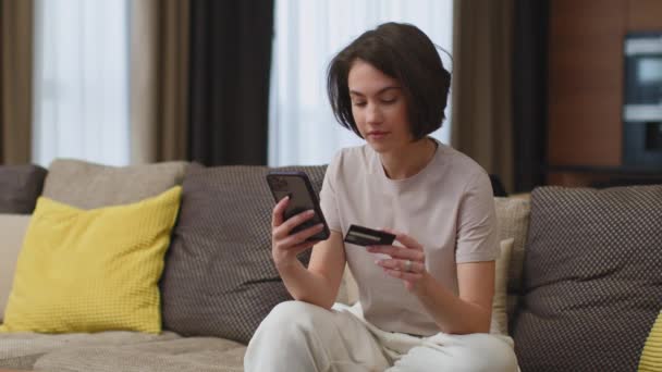 Mladá dívka nakupuje po telefonu s bankovní kreditní kartou, sedí doma na gauči. Online nakupování, e-commerce — Stock video