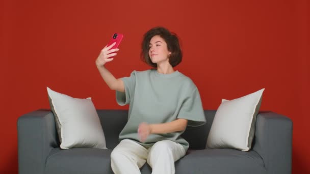 Krásná dívka vezme selfie na svém smartphonu, zatímco sedí doma na gauči. Preen a česat vlasy před kamerou telefonu. — Stock video