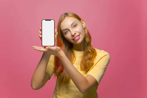 Smiling beautiful girl showing phone with empty blank mock-up screen, advertising new apps on pink studio background — Stock Photo, Image