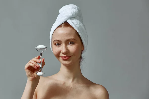 Menina agradável em toalha mostrando massageador jade rolo para rejuvenescimento, pele saudável, rotina diária de cuidados com a pele — Fotografia de Stock