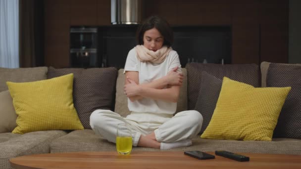 Плохая молодая женщина в шарфе страдает от гриппа, согревается, чувствует холод, замерзает, чихает, сидит дома на диване — стоковое видео