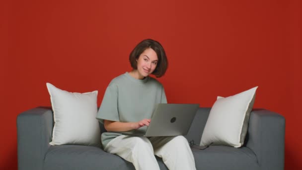 Egy fiatal nő otthon dolgozik egy laptopon. Távoli iroda. Felteszi a szemüveget, hogy munkamenetet kezdjen a kanapén.. — Stock videók