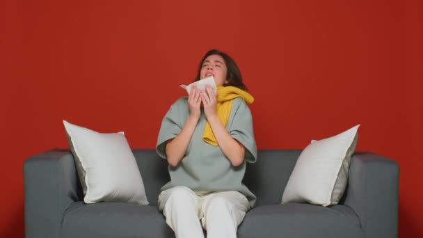 Dość chora młoda dziewczyna kichająca w serwetce, cierpiąca na biegnący nos, objawy grypy, przeziębienie sezonowe, siedząca na kanapie — Wideo stockowe