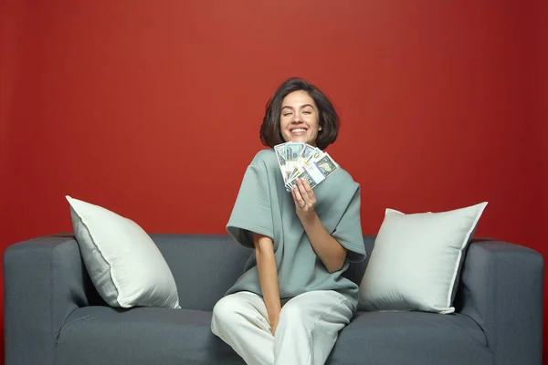 Úspěšná bohatá mladá dívka držící peníze peníze s úsměvem, ukazující bankovky vychutnat finanční úspěch, sedí na pohovce — Stock fotografie