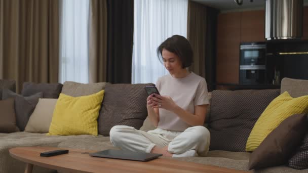 Šťastná mladá dívka s telefonem pomocí aplikace sociální sítě chatování s přáteli nebo přítelem sedí na pohovce doma — Stock video