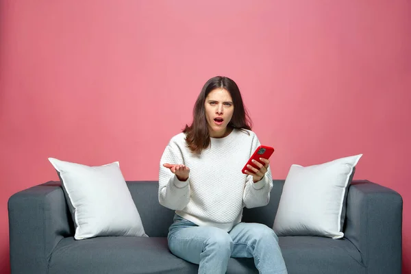 Shocked annoyed young woman holds phone irritated by by bad news or spam in message online, sitting on sofa — Stock Photo, Image