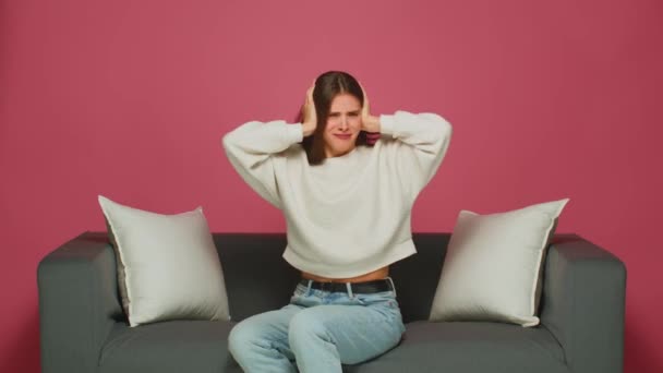 Wkurzona młoda dziewczyna wkłada palce do uszu nie słuchając, odmawia słuchania hałaśliwych sąsiadów, siedzi na kanapie w domu — Wideo stockowe