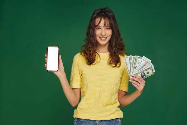 Šťastná mladá dívka držet peníze v ruce a telefon s prázdnou šablonou obrazovky, inzerovat bankovní aplikace na zeleném pozadí — Stock fotografie