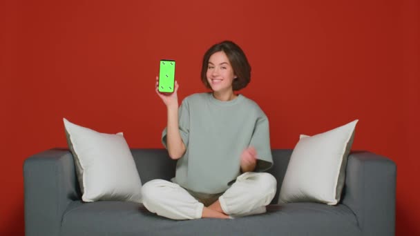 Eine junge Frau, die zu Hause auf der Couch sitzt, zeigt einen grünen Smartphone-Bildschirm. Werbung für die Anwendung. Lächeln und gute Laune. — Stockvideo
