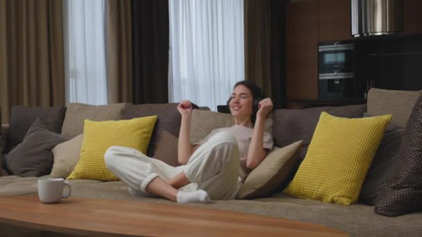 Glückliches junges Mädchen setzt moderne drahtlose Kopfhörer auf den Kopf, lügt sich zum Entspannen auf ein gemütliches Sofa und genießt die Lieblingsmusik — Stockvideo